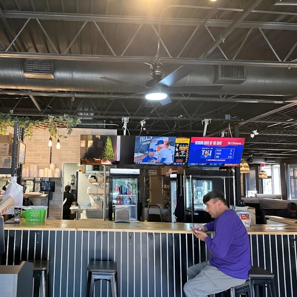 Man in purple shirt sitting at a bar, looking at his phone, with TVs showing sports in the background.